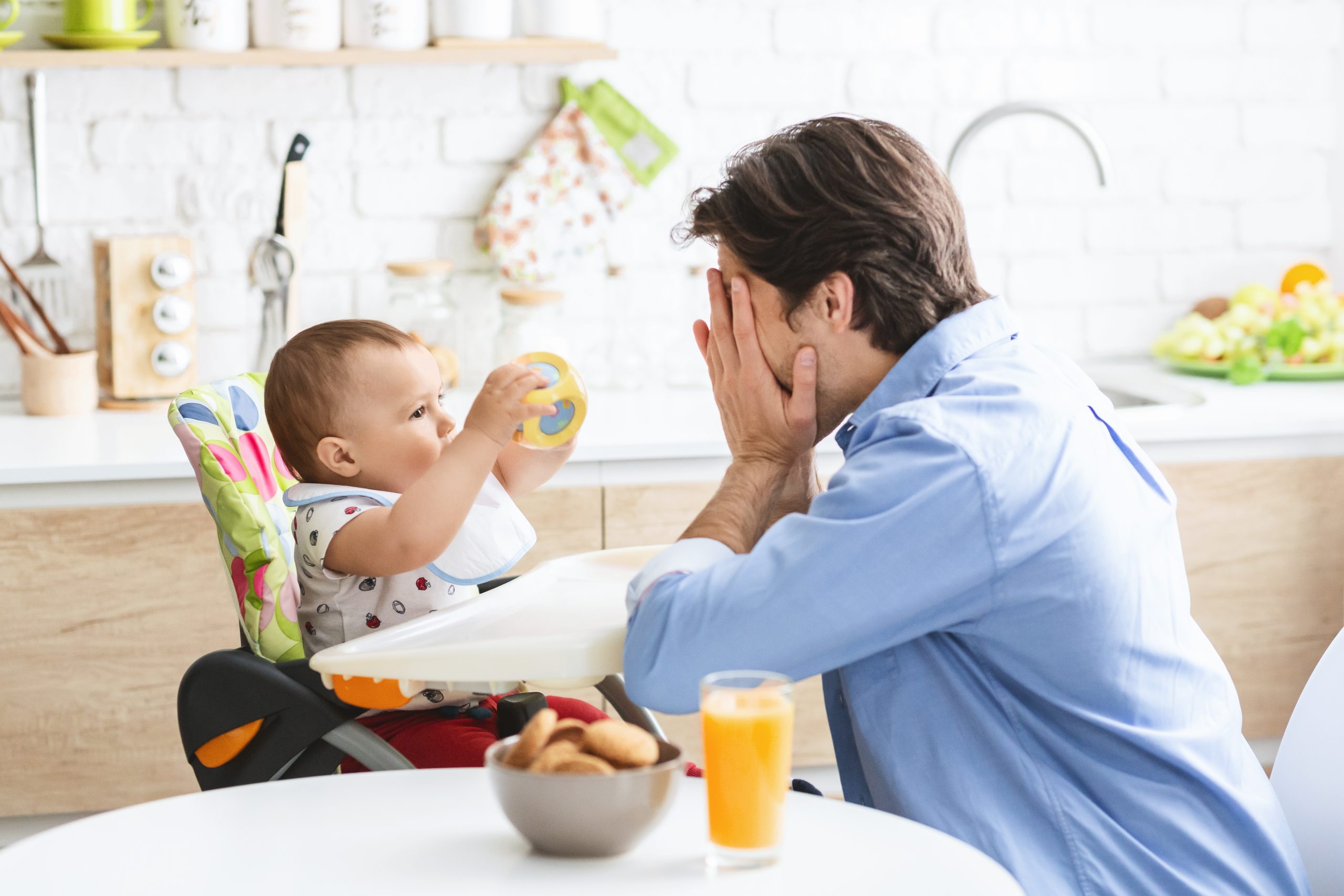 Cheerful father playing peekaboo with his baby son in a kitchen setting.