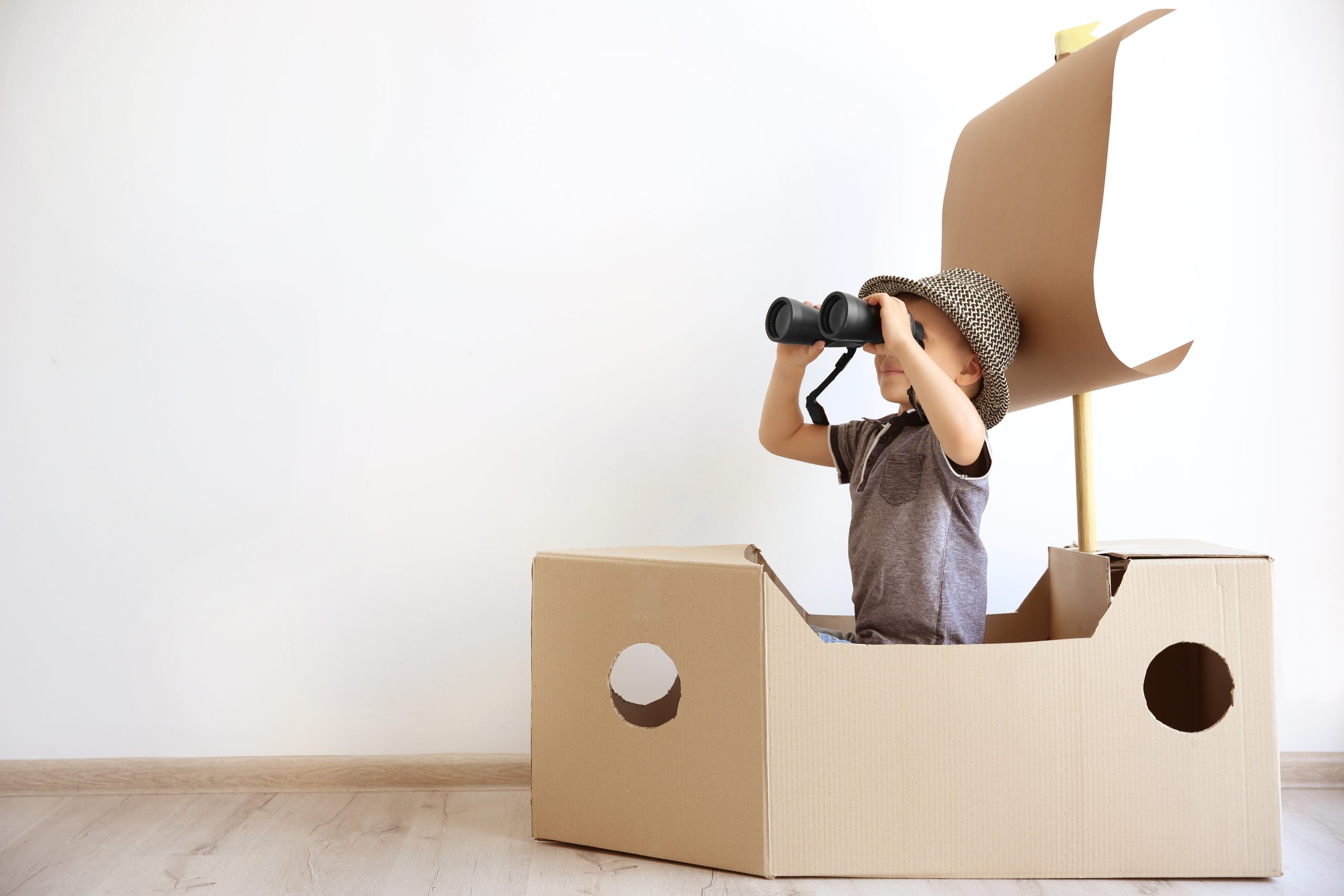 Little boy joyfully interacting with a cardboard ship against a white wall.