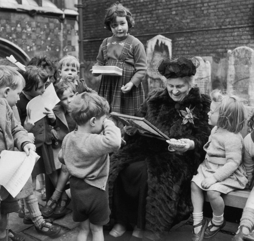 A vintage black-and-white photograph of Maria Montessori engaged in teaching a diverse group of children gathered around her.