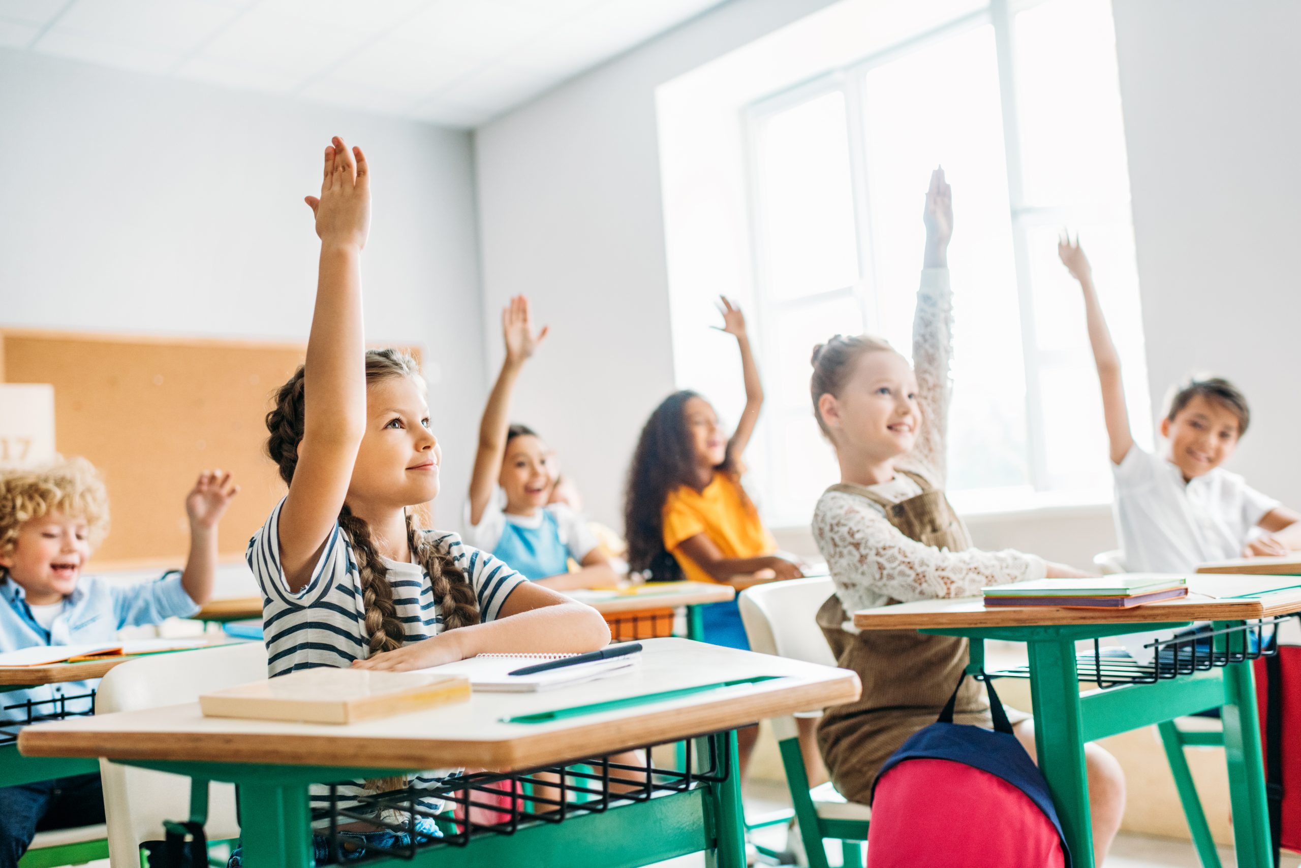 Group of diverse schoolchildren enthusiastically raising their hands to answer a question in class.