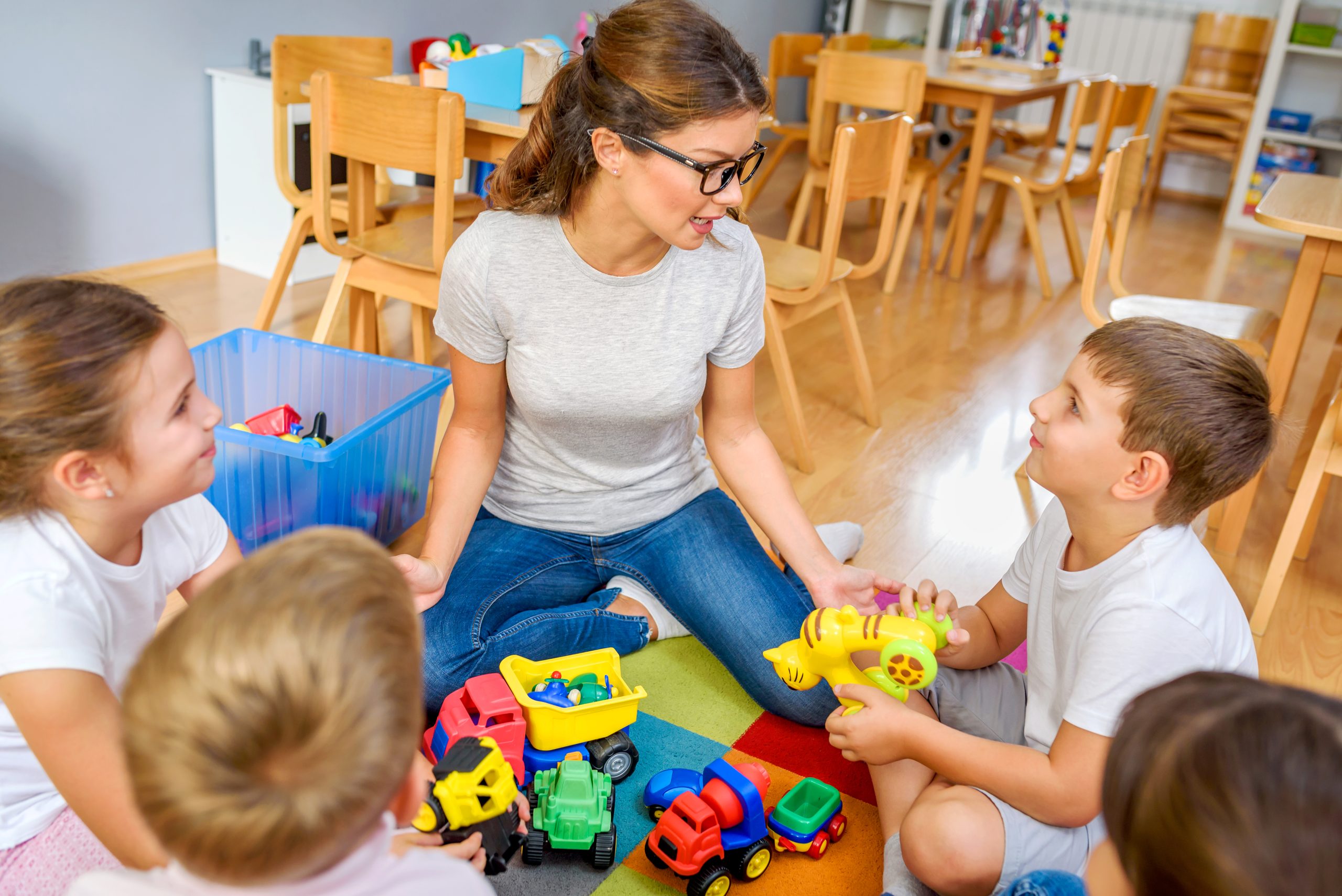 Preschool teacher engaging with a group of attentive children sitting cross-legged on a classroom floor.