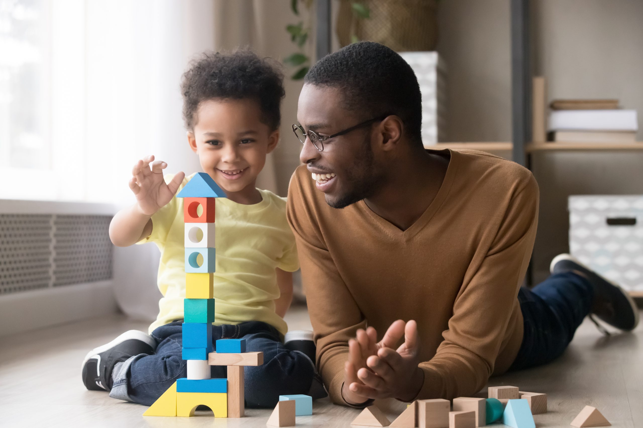 African father and his young son joyfully building a tower together using multicolored wooden blocks on a warm floor at home.