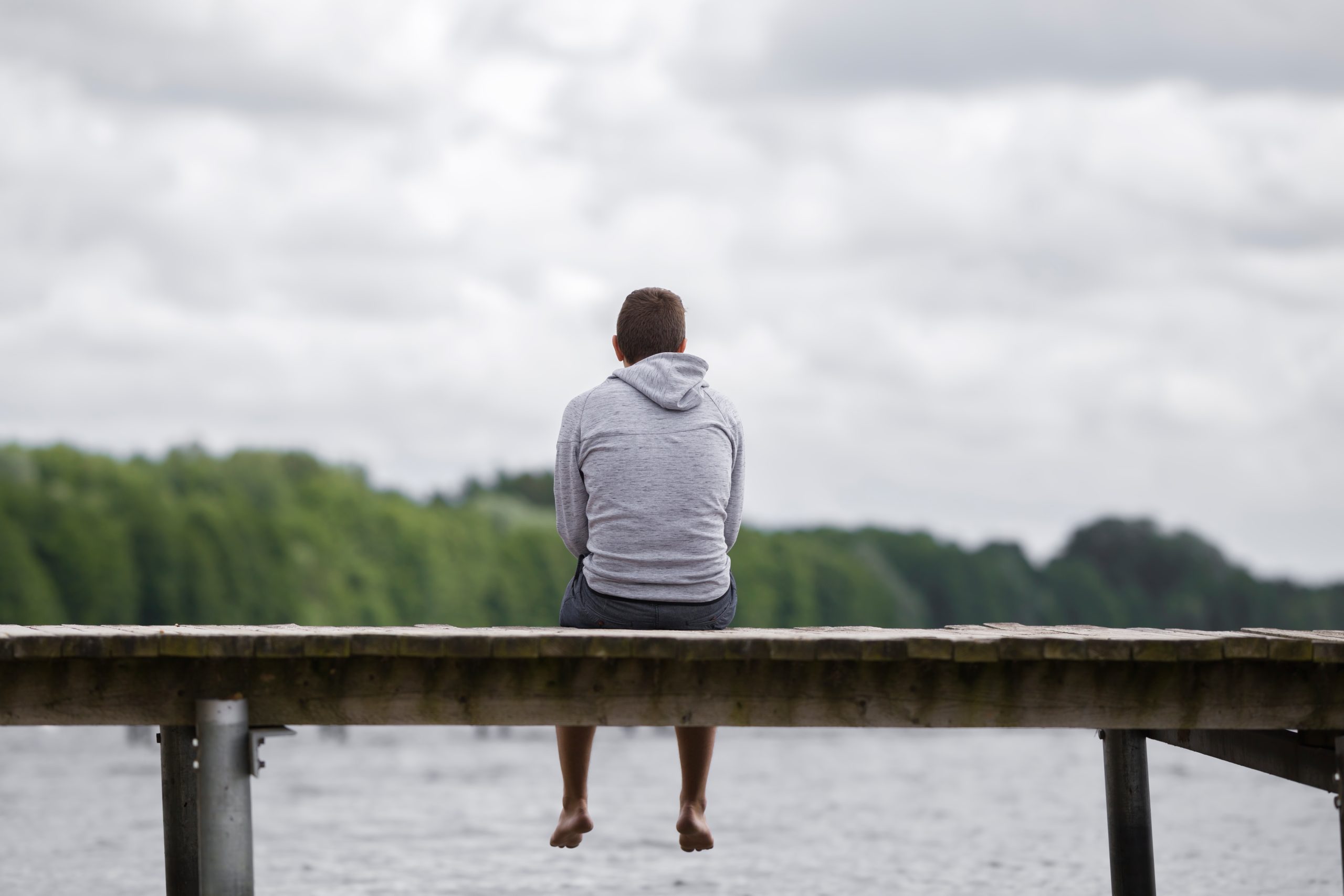 Young man sitting alone on the edge of a footbridge, gazing thoughtfully at a serene lake surrounded by green trees under a cloudy summer sky.