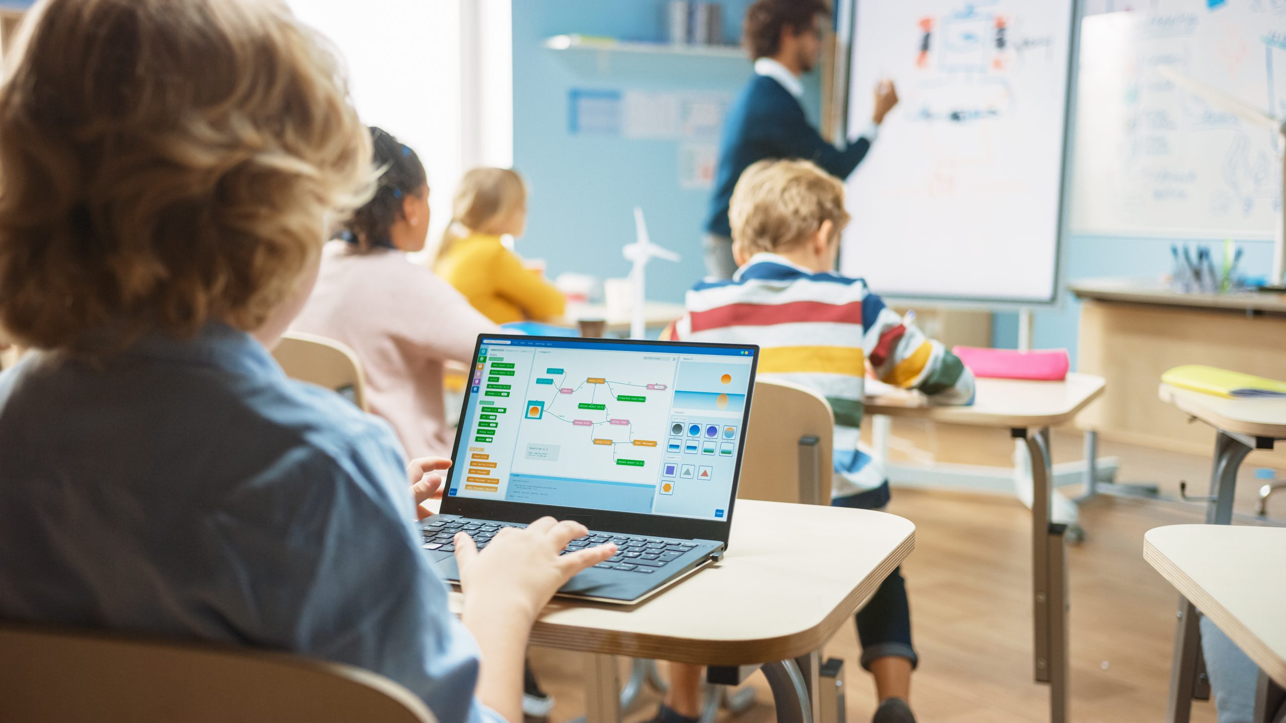 Over-the-shoulder view of a young boy using a laptop with programming software in an elementary school science class, while the physics teacher explains the lesson to a diverse group of students.