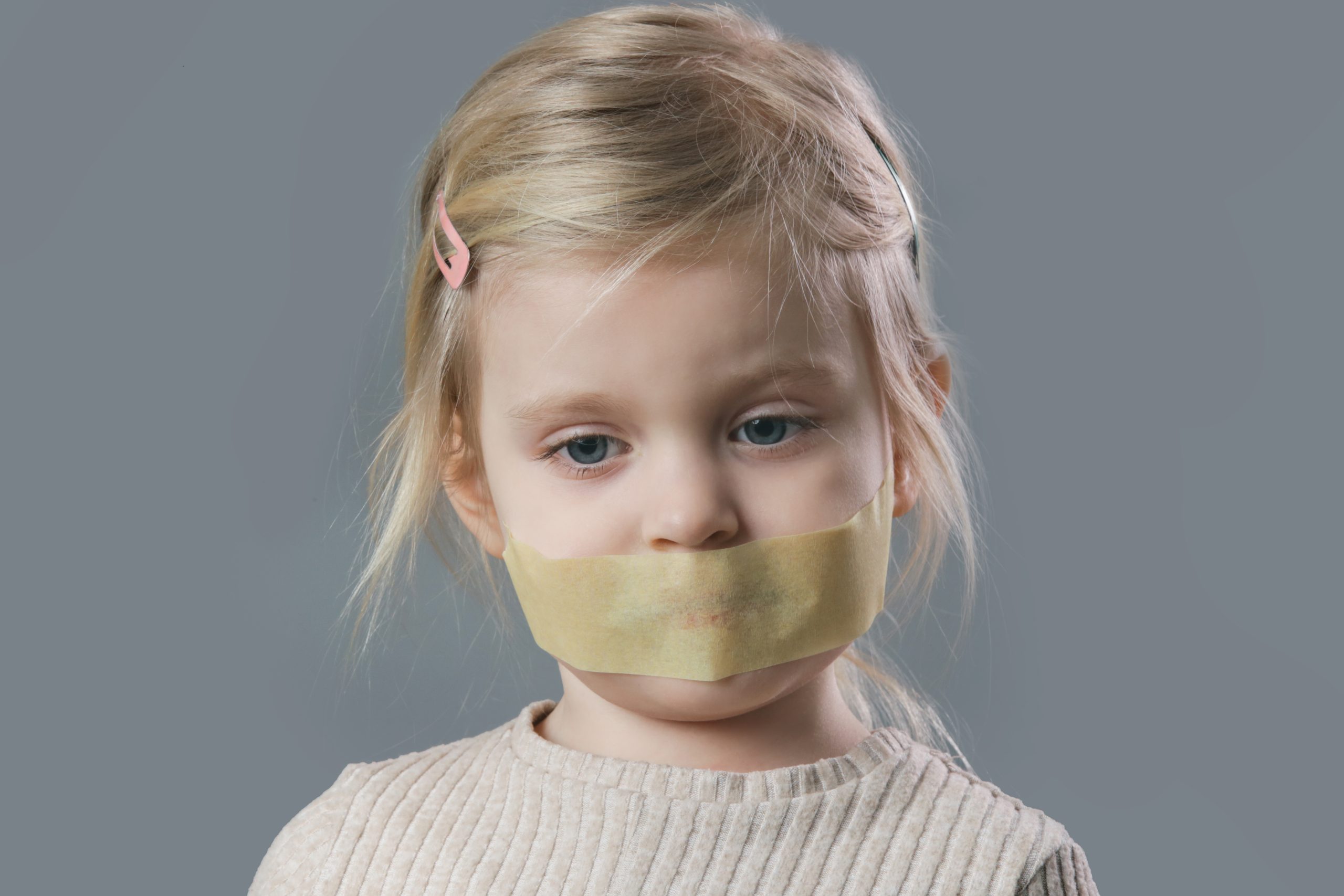 Studio portrait of little girl with adhesive tape over her mouth, symbolizing restricted freedom of speech.