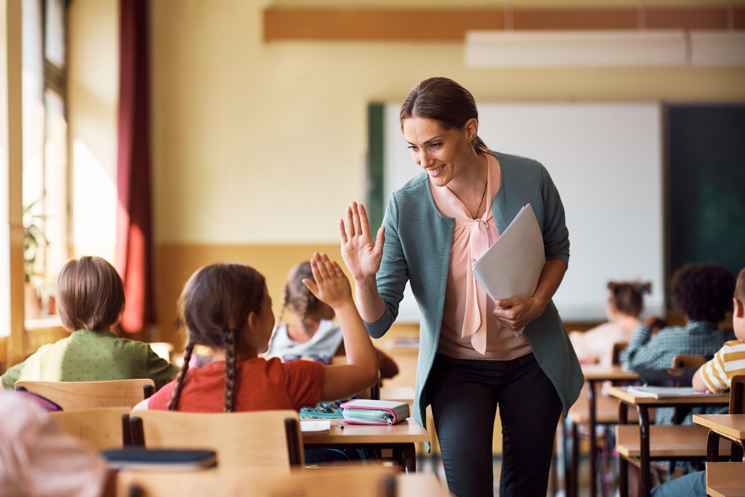 Smiling elementary school teacher giving high-five to a student in the classroom.