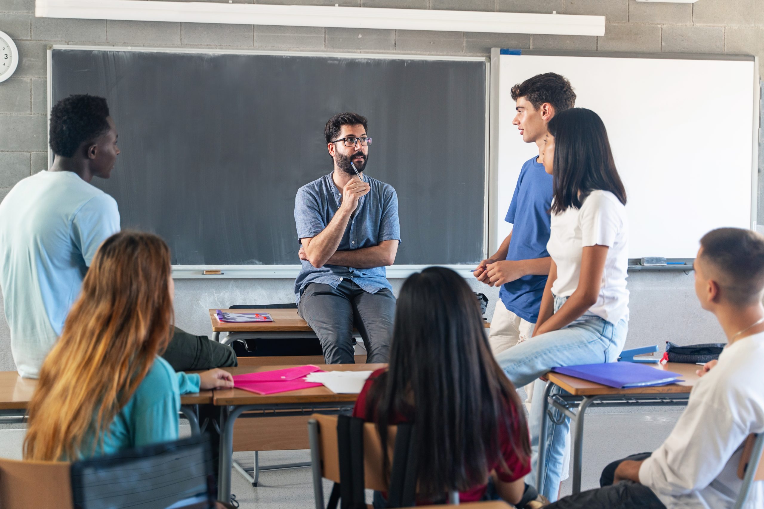 Group of teenagers and a young male teacher sitting in a classroom, engaged in discussion.