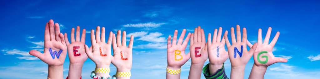 Children's hands arranging letters to form the words 'Well Being' against a bright blue sky backdrop.