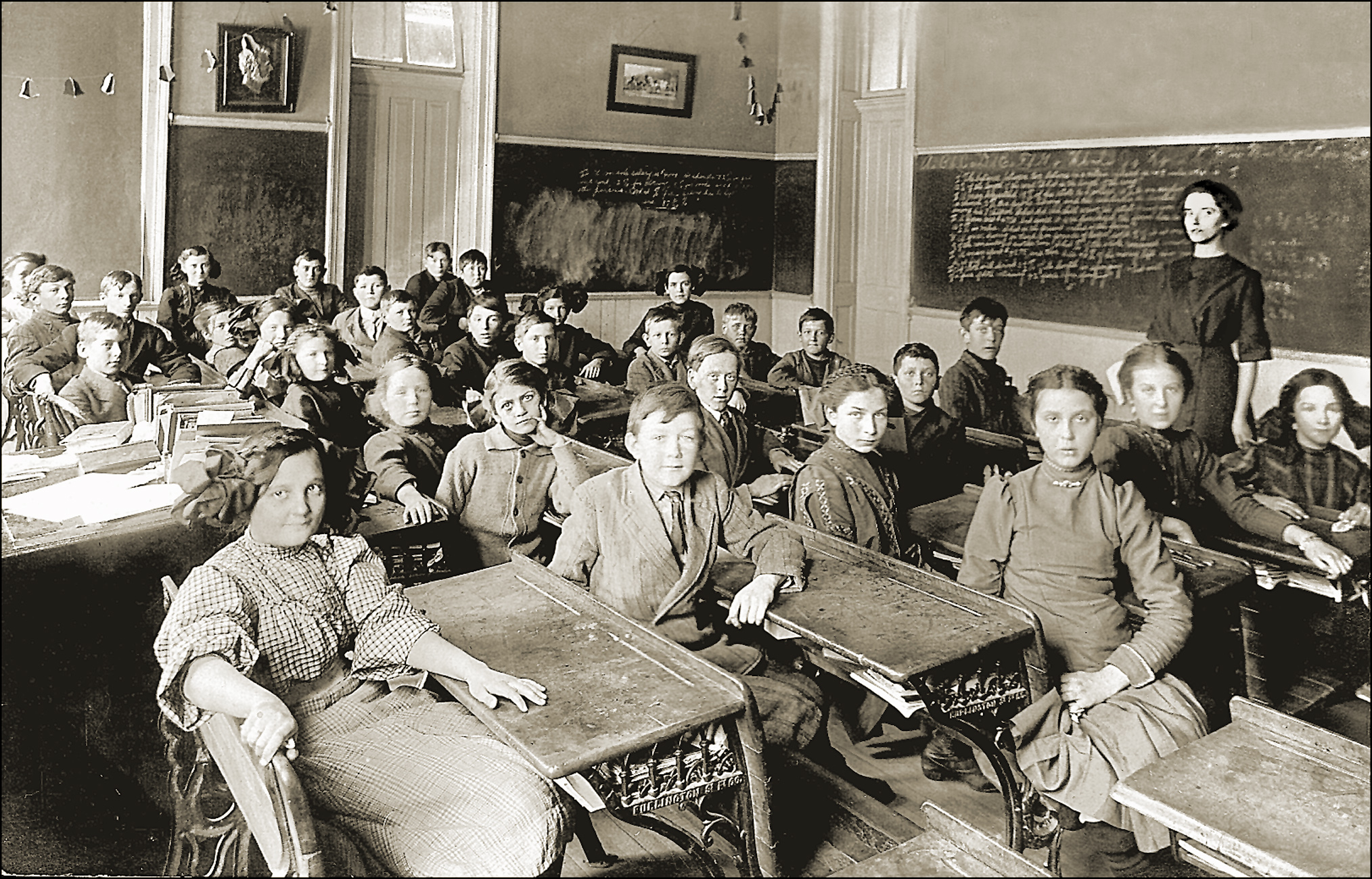 Black-and-white vintage photo of a classroom with students seated at wooden desks, and a teacher standing near a chalkboard