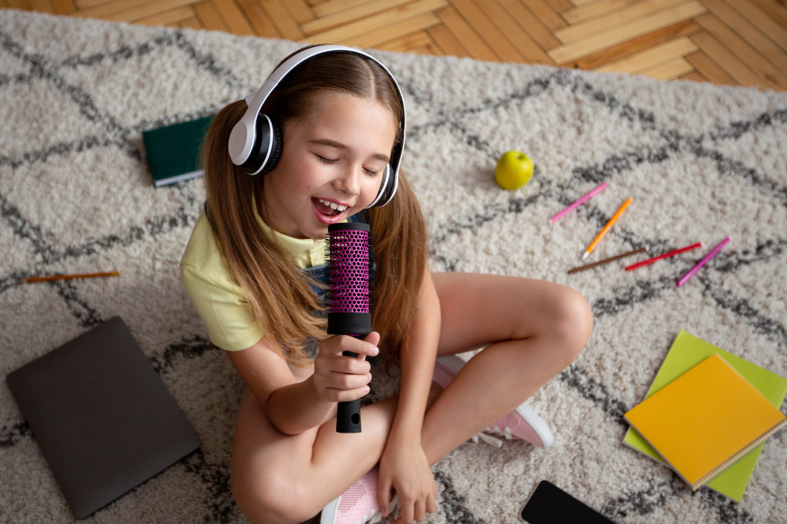 High-angle view of a teenage girl sitting on the floor, singing into a hairbrush as if it's a microphone while wearing headphones.