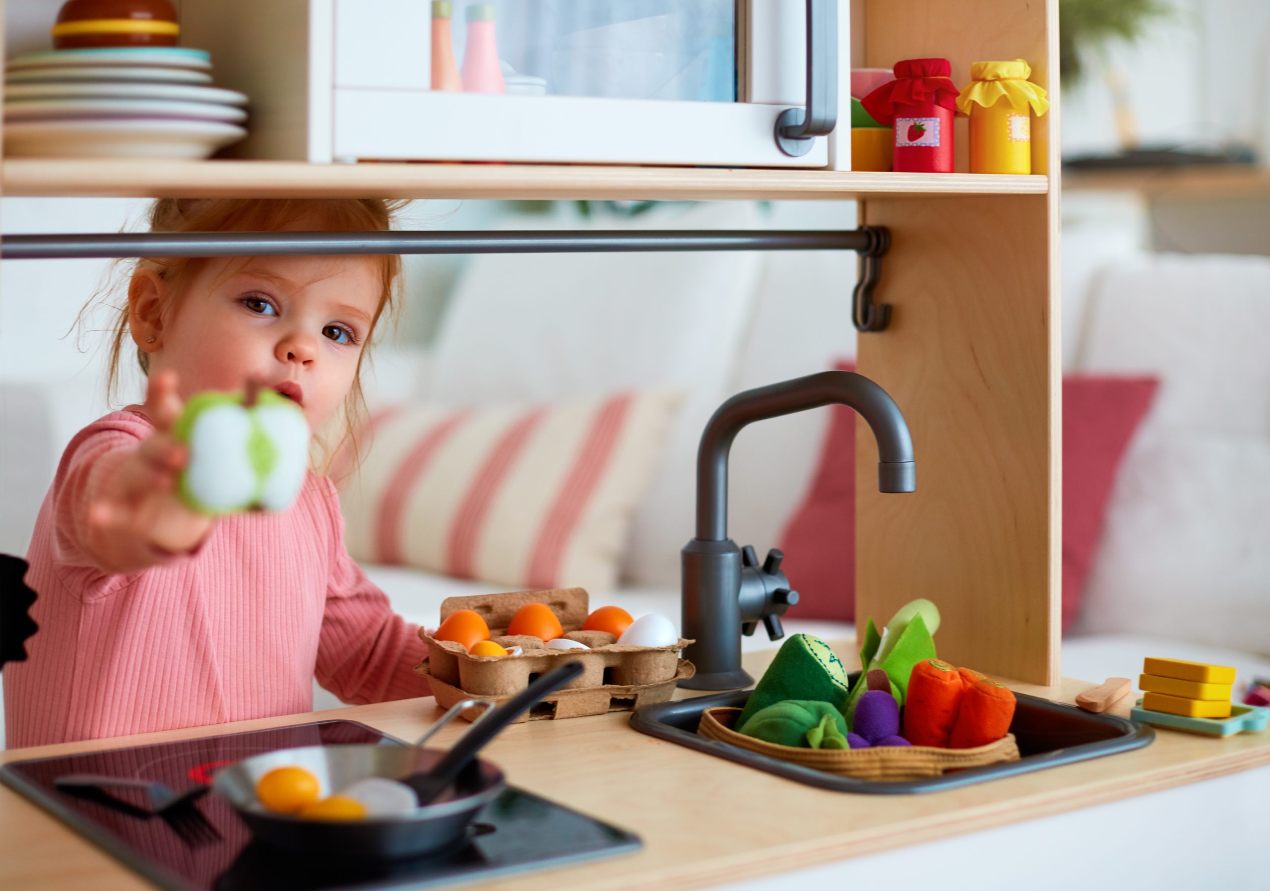 Toddler girl playing with a toy kitchen, preparing eggs and offering a slice of apple.