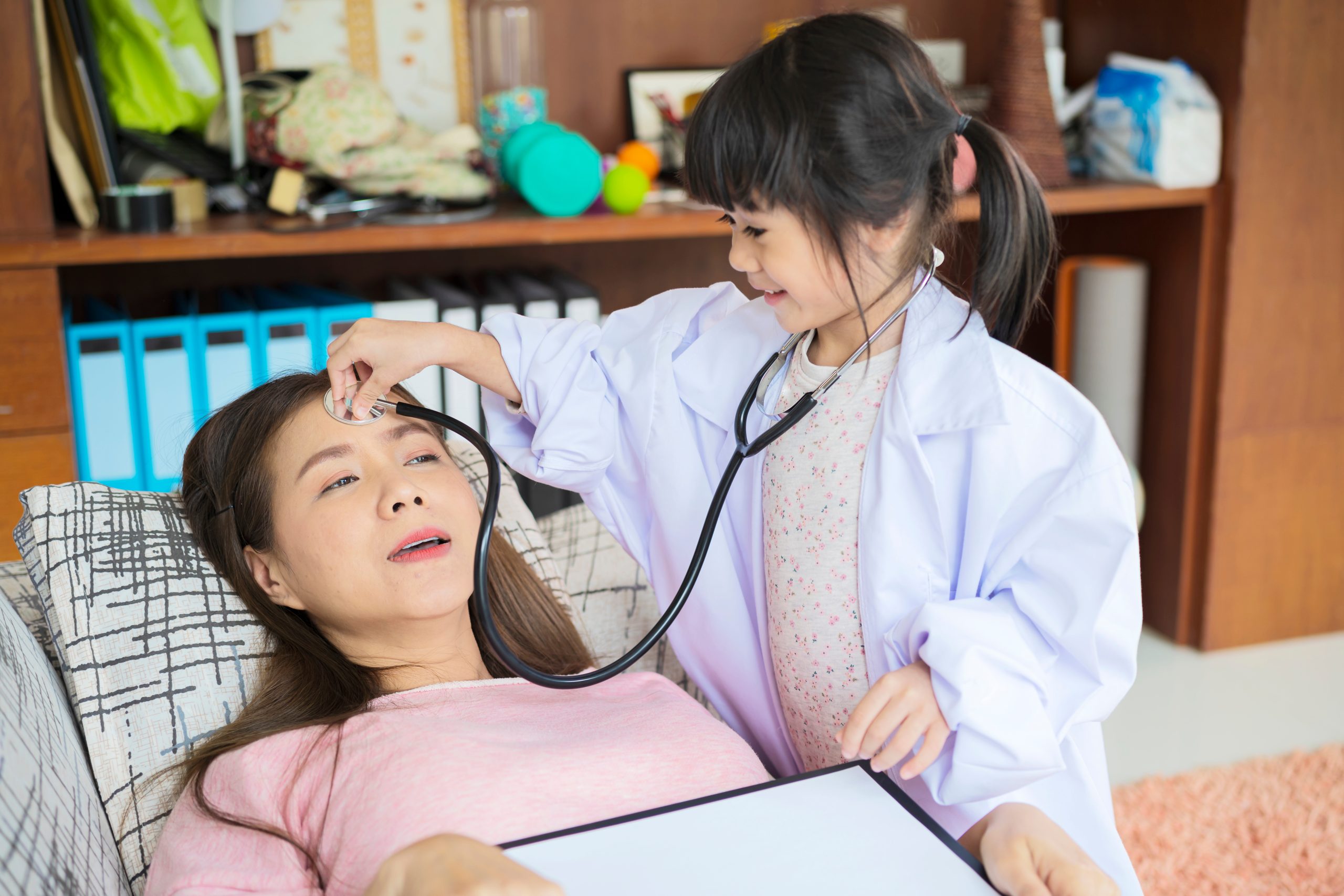 Cute little Asian girl pretending to be a nurse, using a stethoscope to listen to her mom's heart at home.