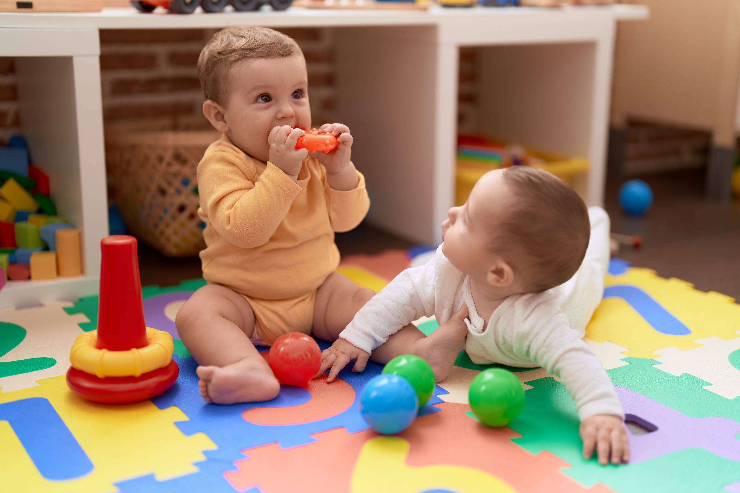 Two toddlers engaged in play with toys while seated on the floor of a kindergarten.