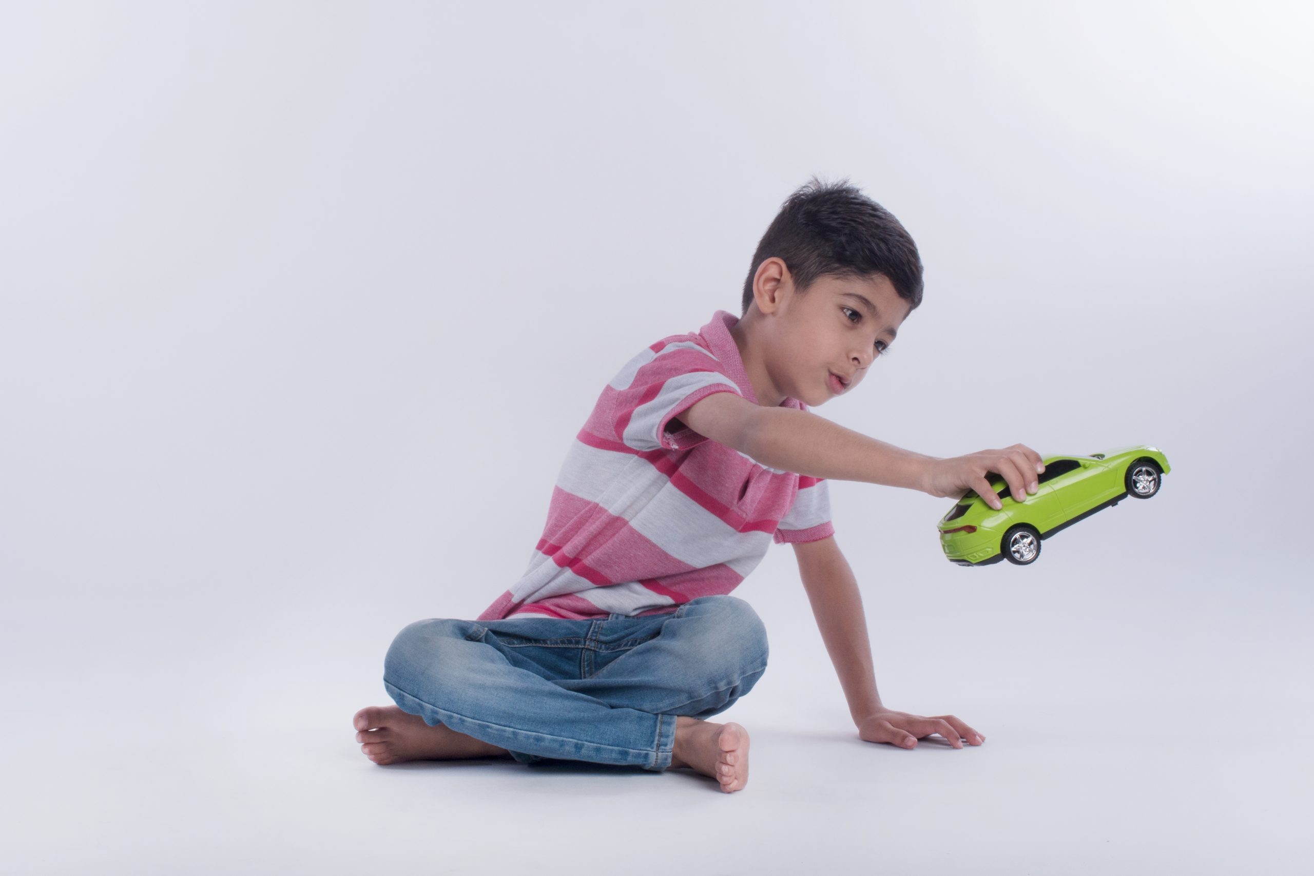 A happy young boy sitting on the floor, engrossed in playing with a green toy car against a white background