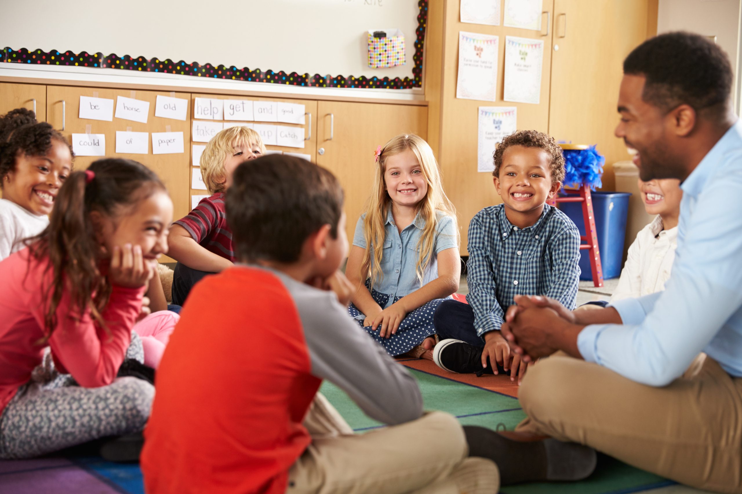 Group of elementary school children sitting cross-legged, actively listening to one another with a teacher guiding the discussion.